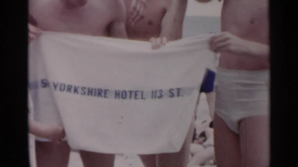 Home-movie frame from a 1954 New York beach showing beachgoers holding a white towel with partial Yorkshire Hotel text.