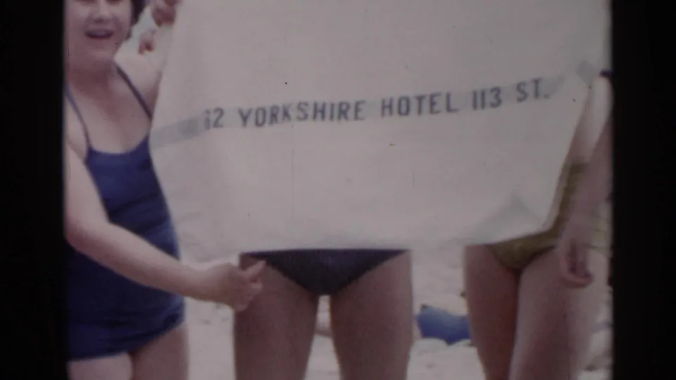 A woman in a dark swimsuit and young men at a New York beach hold a white towel with Yorkshire Hotel and 113 St. lettering.