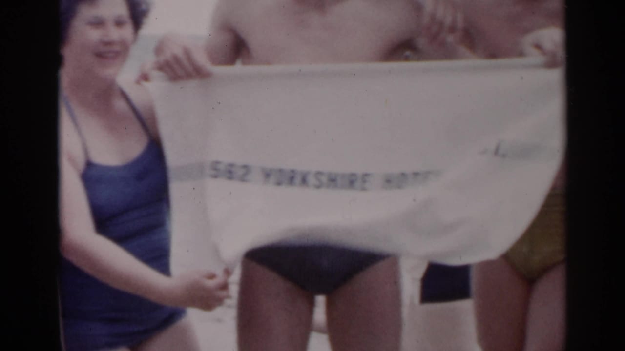Adults in swimsuits hold a white towel with partly readable text on a beach.