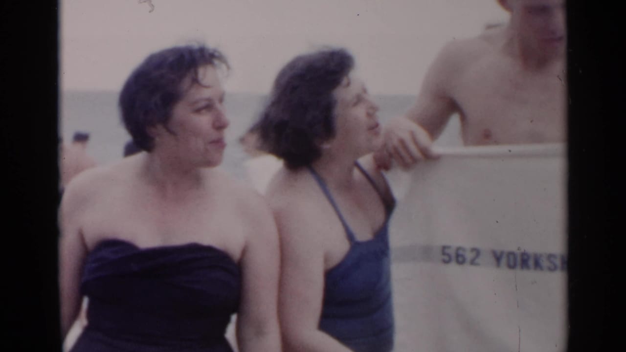 1954 New York beach frame with two women in blue swimsuits near a white towel and ocean background.