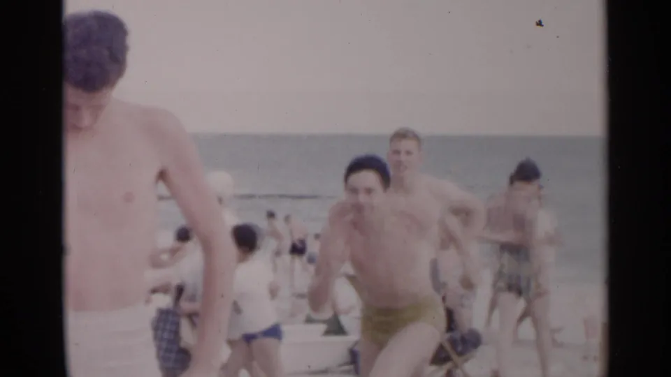 1954 New York beach home-movie frame showing men in trunks moving across sand with ocean and distant bathers behind.