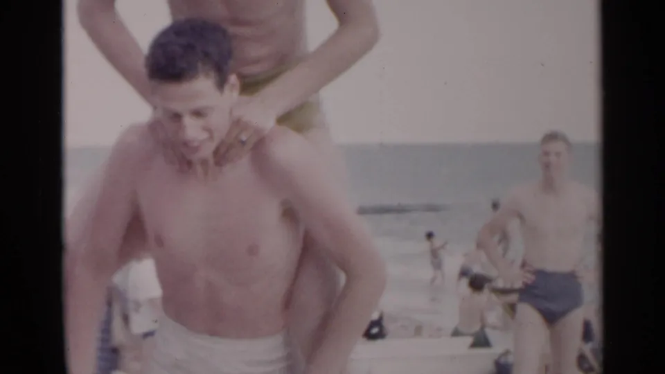 1954 New York beach frame showing a smiling man carrying another person on his shoulders with bathers and ocean in back.