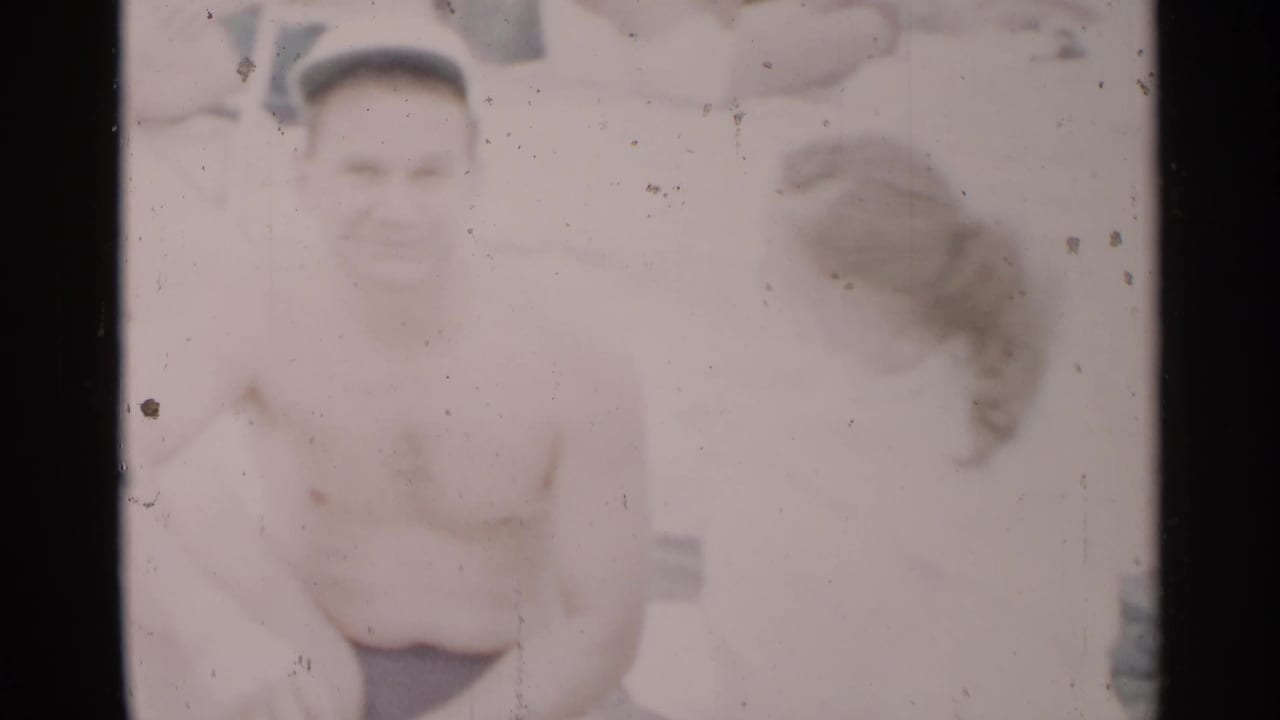 In 1954 New York beach footage, a shirtless man in dark swim shorts and a white cap sits near a woman with her back partly to camera.