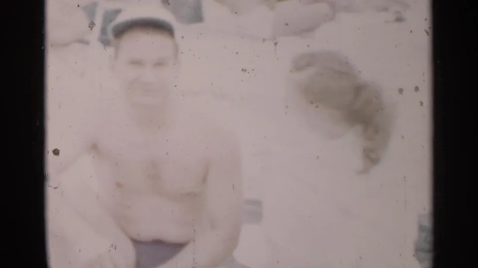 In 1954 New York beach footage, a shirtless man in dark swim shorts and a white cap sits near a woman with her back partly to camera.