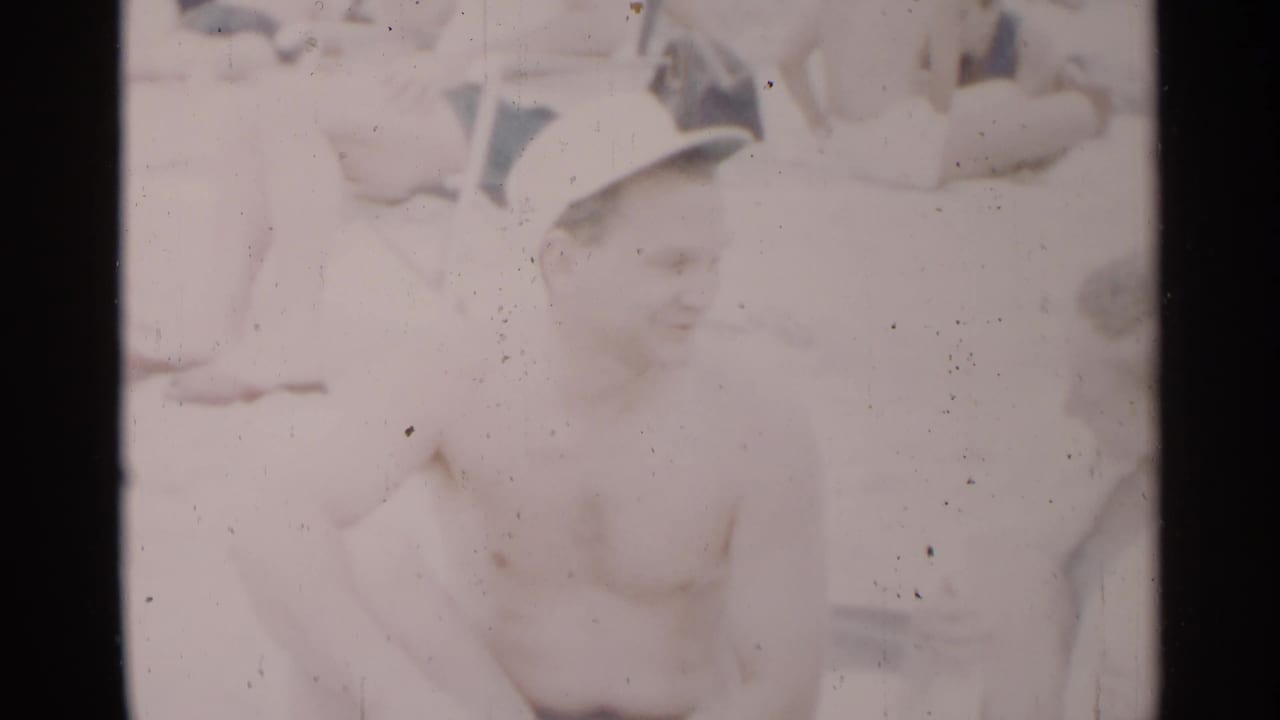 Smiling shirtless man in a white cap sits on the beach with people behind him.