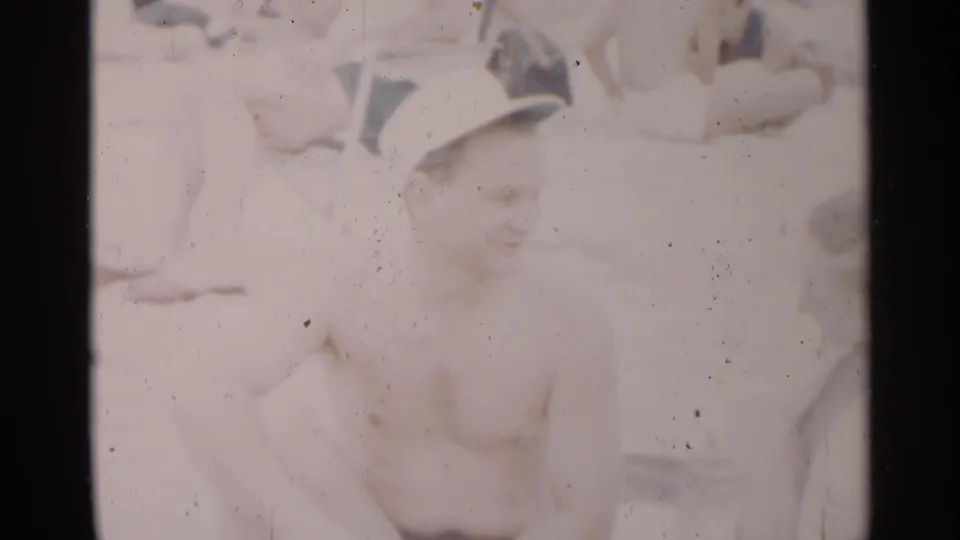 The man in a white cap smiles toward the camera in faded 1954 New York beach footage, with other beachgoers partly visible behind him.