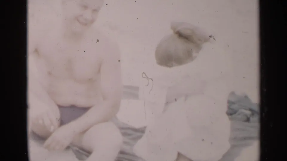 A shirtless man sits in the foreground as a woman with a bun turns away, captured in soft, overexposed 8mm beach footage.
