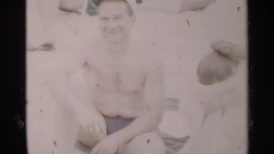 At the end of the clip, the shirtless man smiles from a seated beach pose while the woman beside him remains turned partly away.