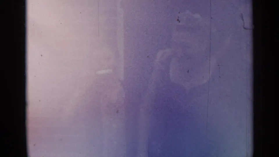 Faint 1954 New York home-movie frame with indistinct adult figures obscured by blue-purple exposure and film edge.