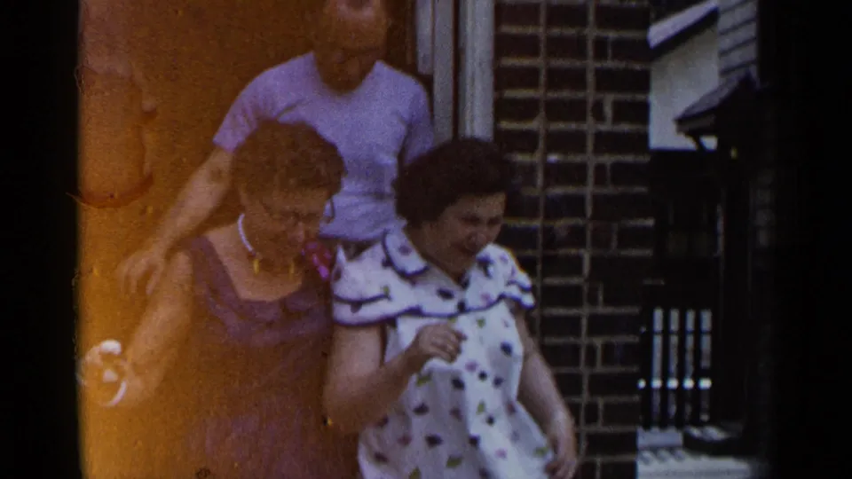 1954 New York home-movie frame with two smiling women and a man close behind them at a brick building entrance.