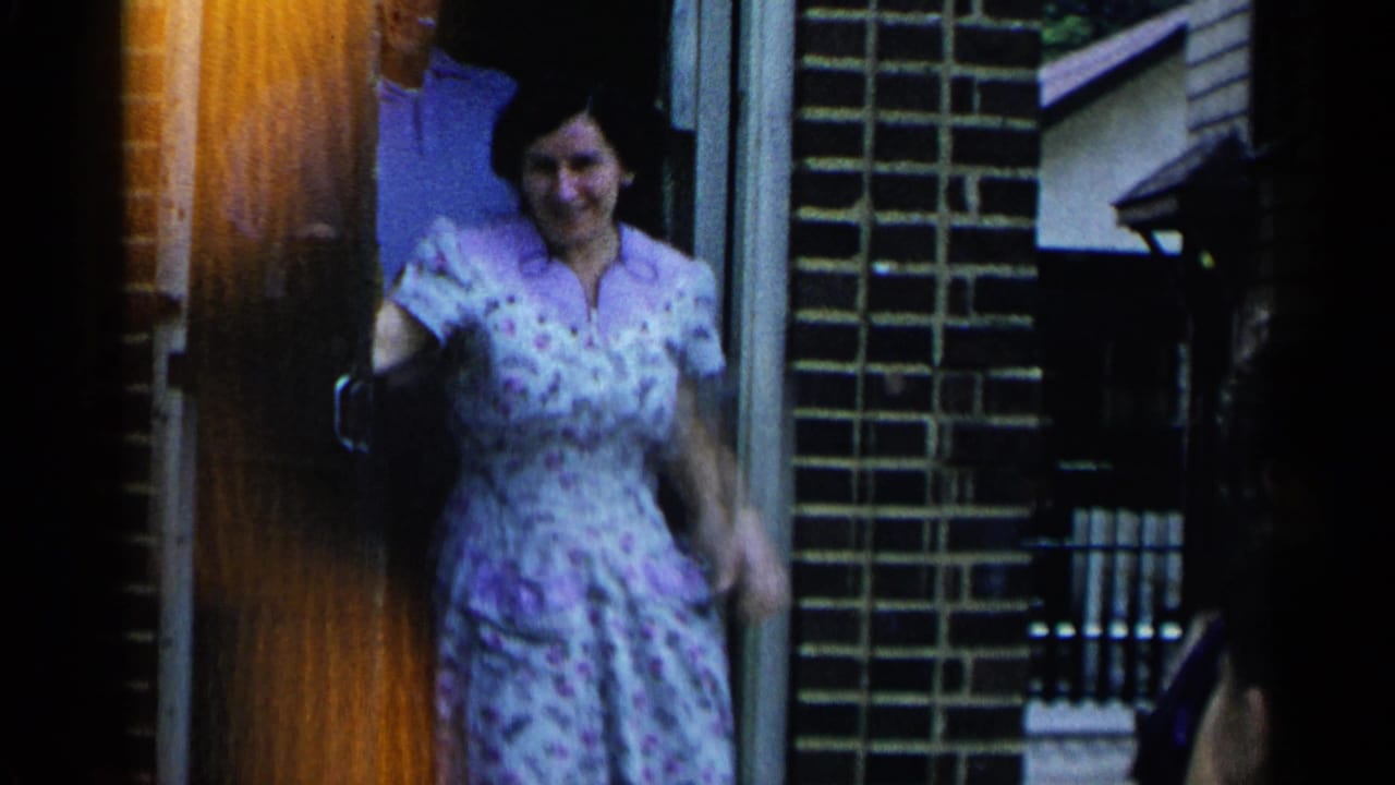 In 1954 New York home-movie footage, a woman in a patterned dress stands at a brick house doorway with another adult partly visible behind her.