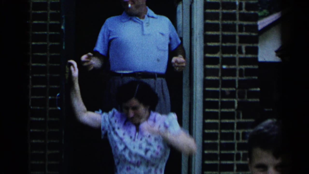 Man in a blue polo stands in a doorway while a woman waves in front of him.