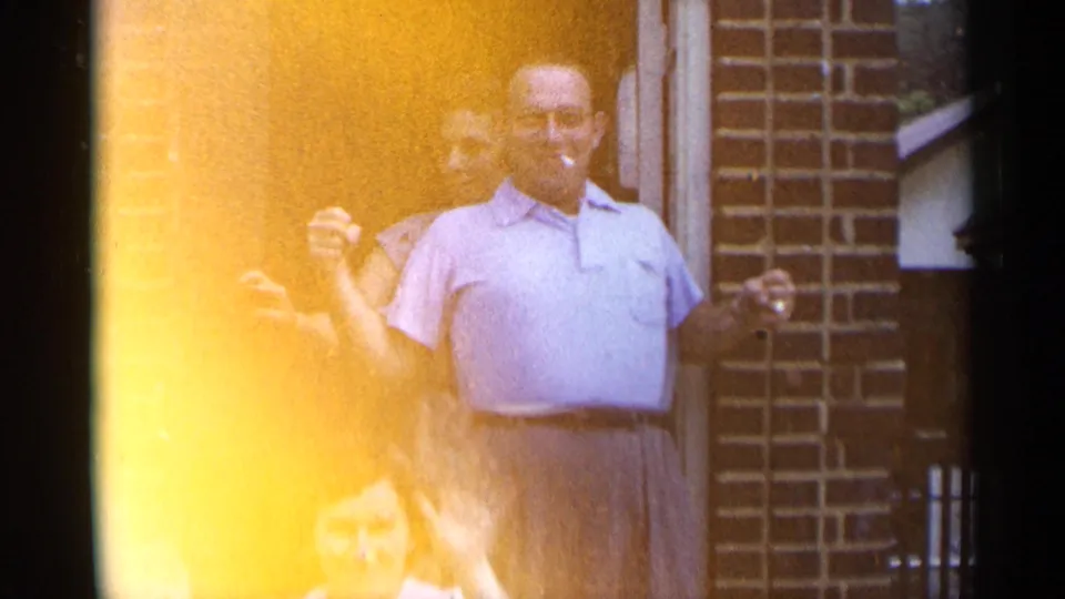 A smiling man with a cigarette stands outside the brick doorway, with two women partly visible around him in the home-movie frame.