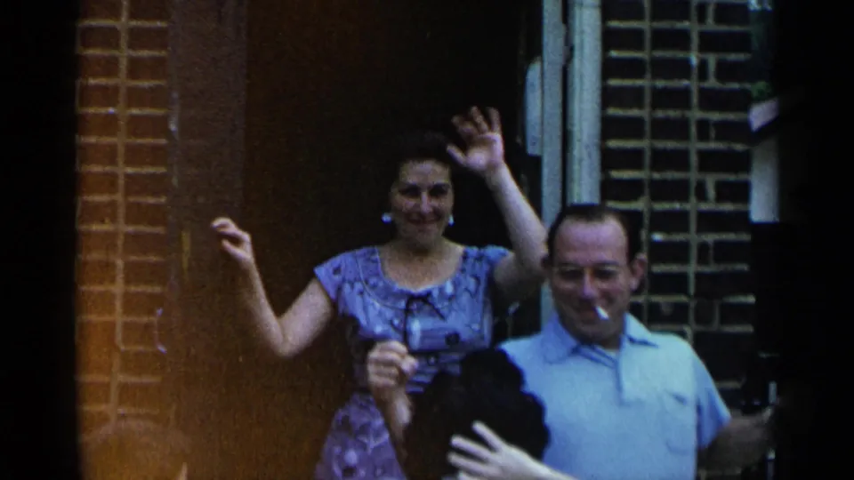 A woman in a patterned dress waves beside a man with a cigarette outside a brick house doorway in 1954 New York home-movie footage.
