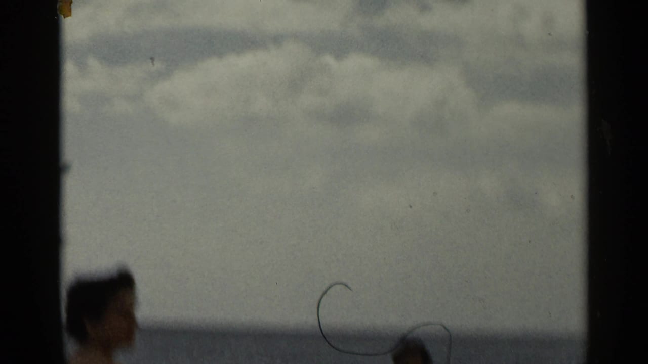 Soft 8mm beach frame with an adult woman near the waterline, open sea and cloudy sky, and dust marks visible on the film.