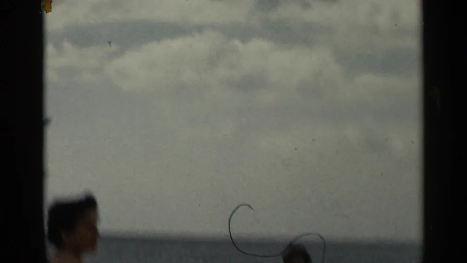 Soft 8mm beach frame with an adult woman near the waterline, open sea and cloudy sky, and dust marks visible on the film.