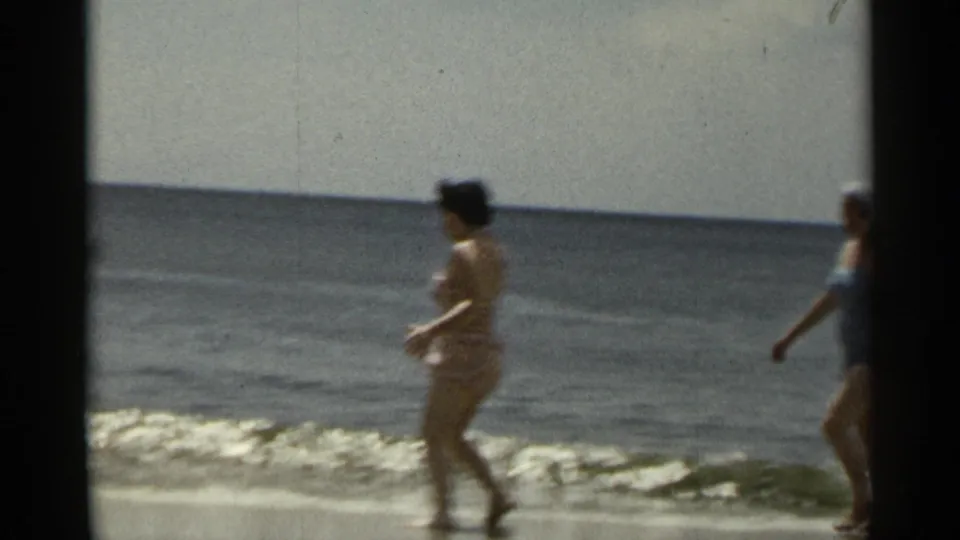 1954 New York beach footage frame showing two adult women in one-piece swimsuits walking through shallow surf under a pale sky.