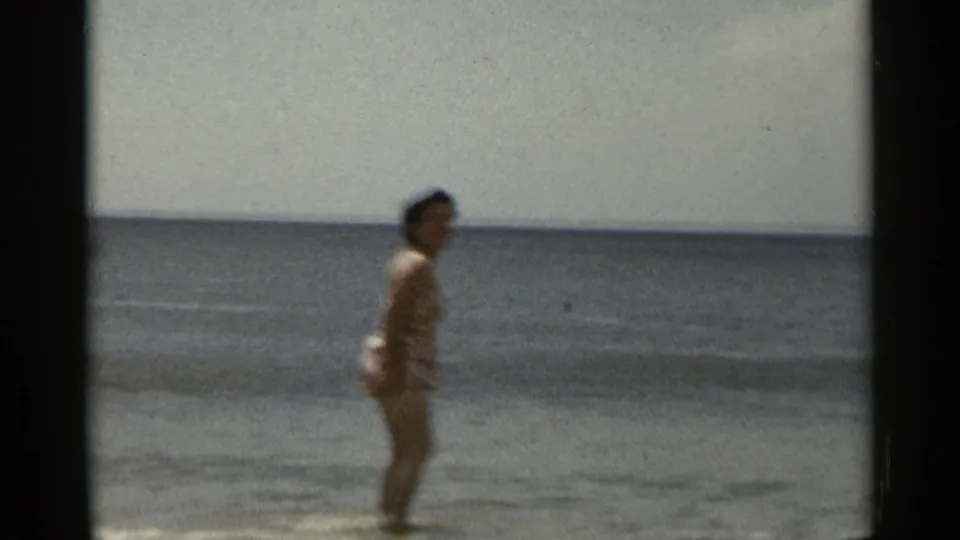 1954 New York beach home-movie frame showing a woman in a swimsuit near gentle waves under a bright sky.