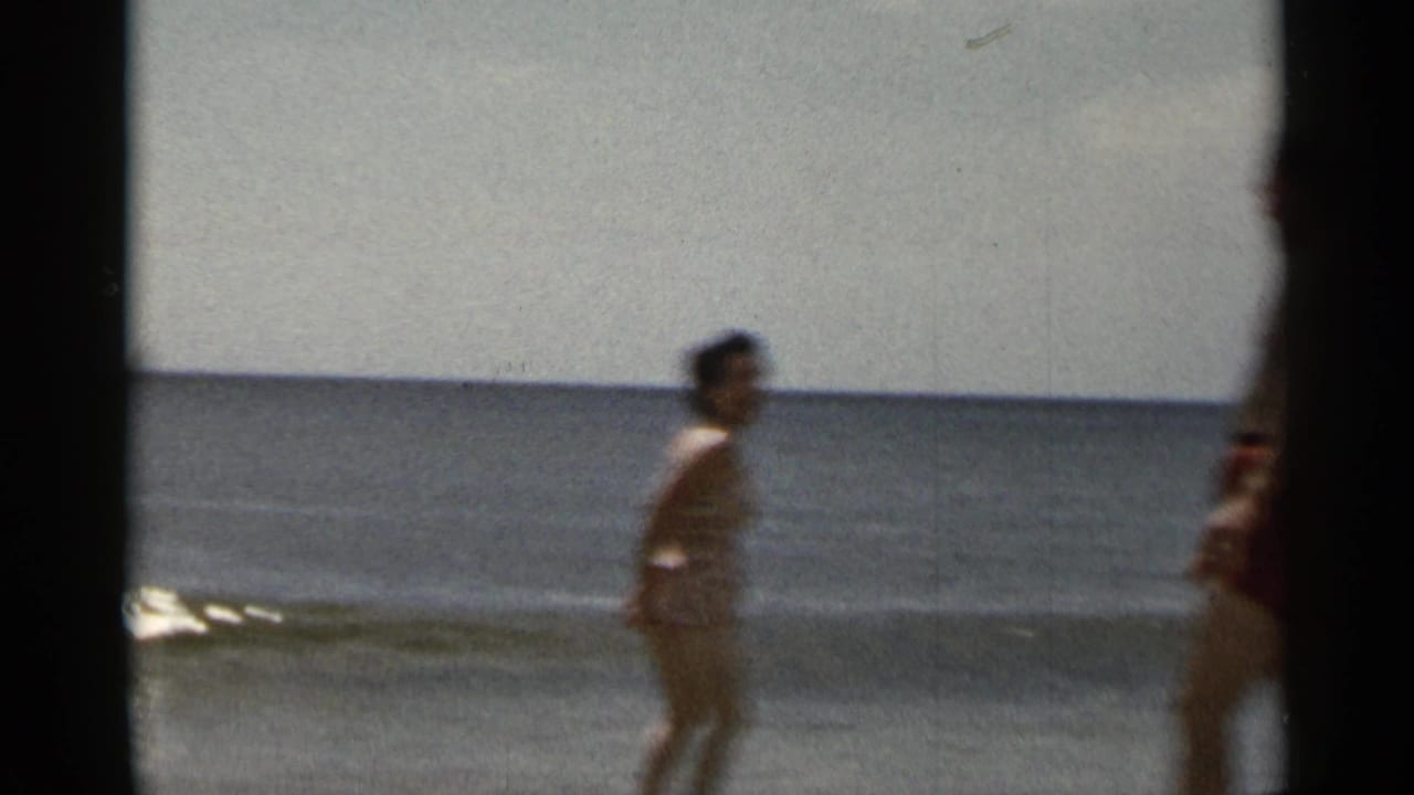 Woman walking along the surf as a man stands partly visible at the right edge.