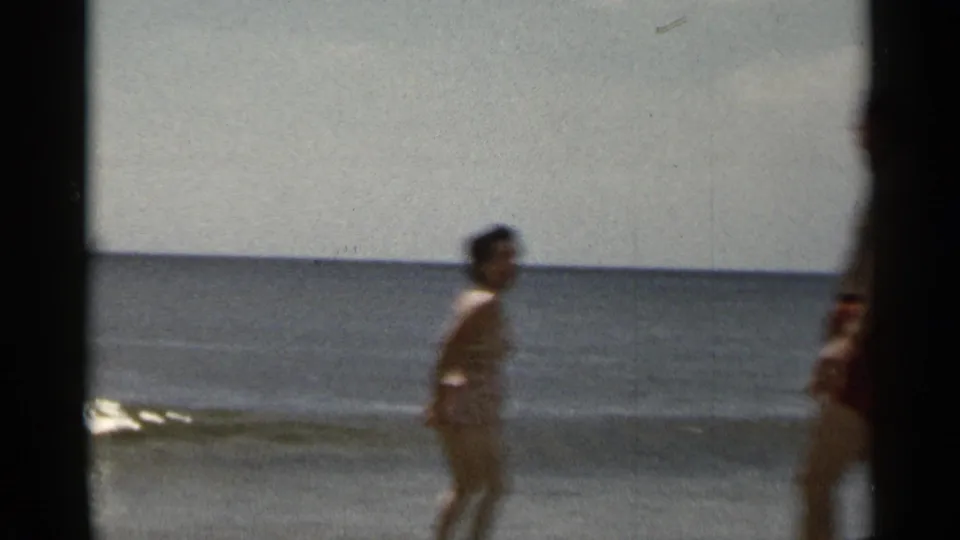1954 New York beach frame with a woman moving through shallow water and a man partly visible near the shoreline.