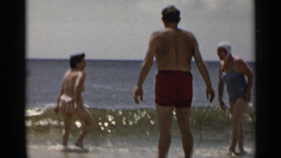 1954 New York beach home-movie frame showing adults in swimsuits gathered at the edge of a breaking wave.
