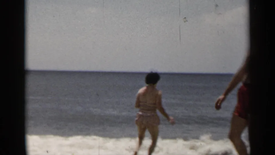 1954 New York beach frame showing a woman near foamy surf while a man in red trunks is cropped at the right edge.