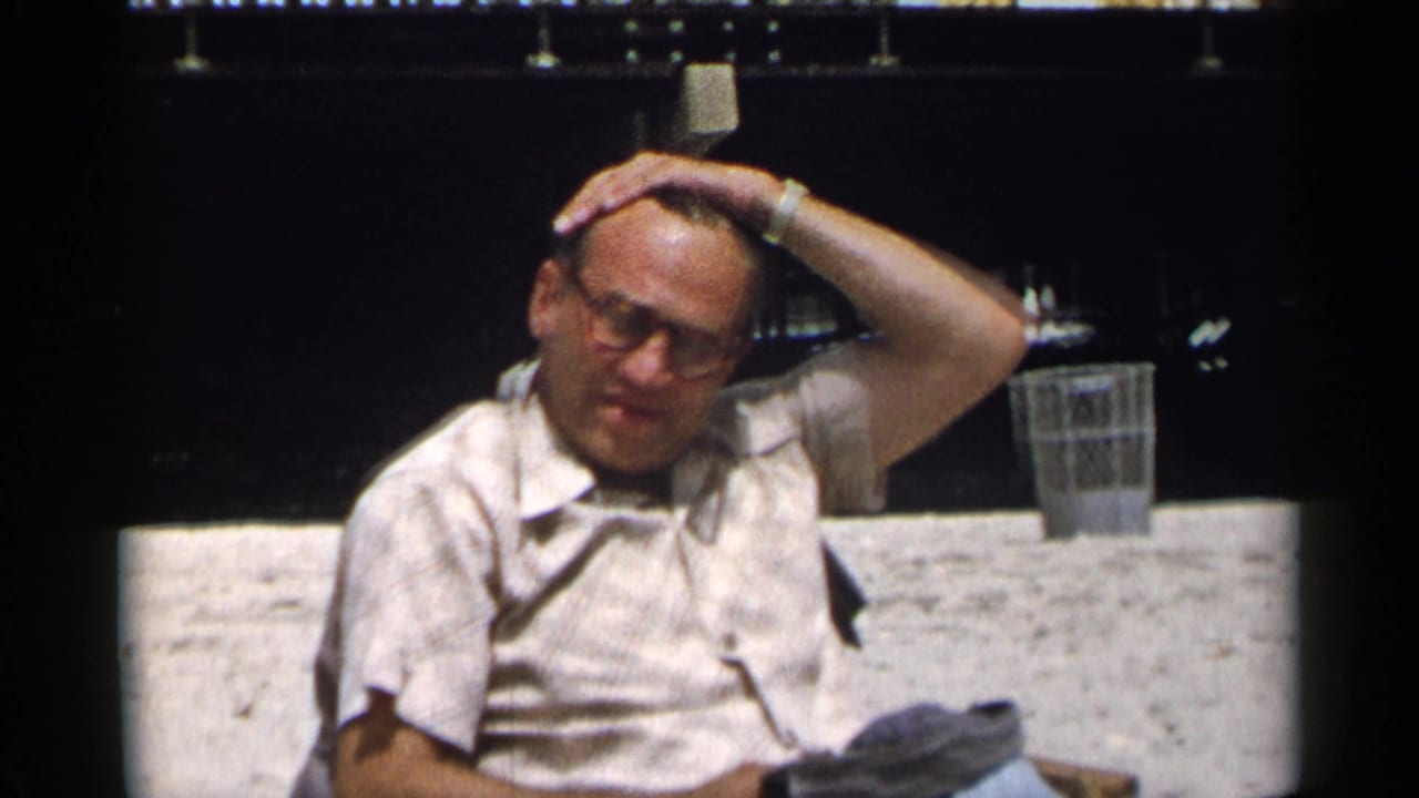 Older man in glasses sits on a bench with one hand on his head.