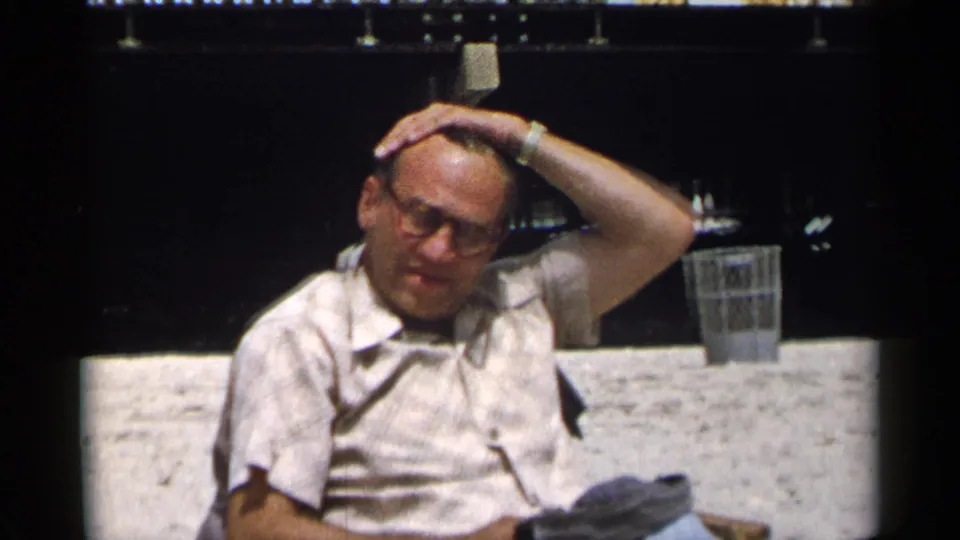In 1954 New York home-movie footage, an older man in glasses sits on a bench and raises one hand to his hair.
