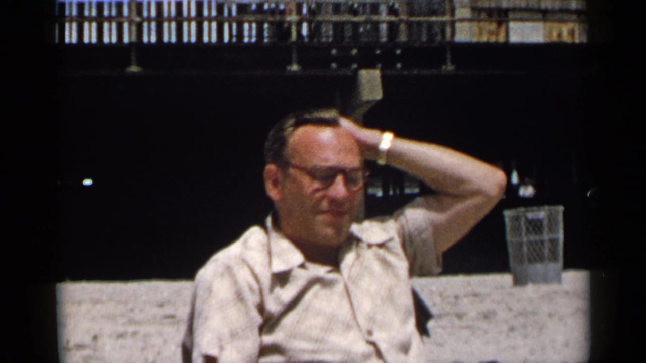 Older man seated on a bench holds one hand behind his head.