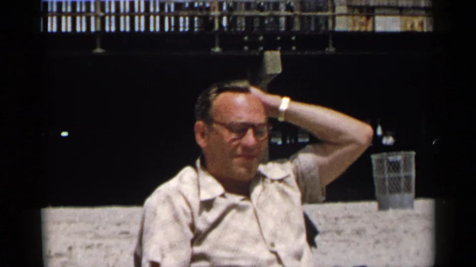 The man remains seated outdoors in New York, wearing glasses and a light shirt as his hand rests behind his head.