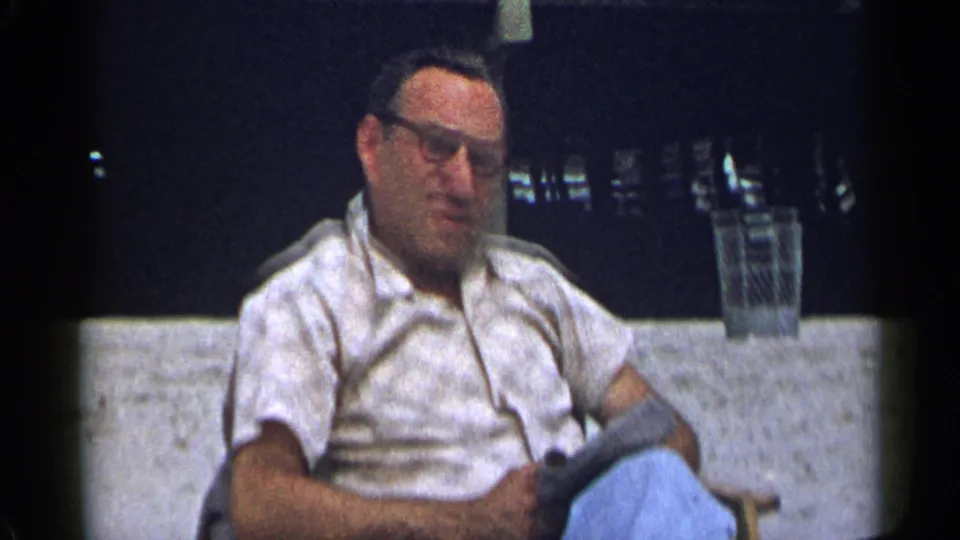The seated man lowers his hand and rests with an item in his lap, with a trash can and railing behind him.