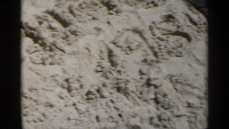 1954 home-movie frame showing a close view of beach sand with footprints and rough surface texture, likely from a New York boardwalk outing.