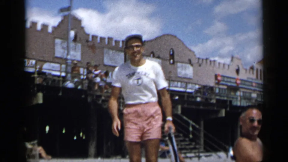 Adult man in glasses and a cap walks toward the camera on a New York boardwalk in 1954, with a crenellated building and beachgoers behind him.