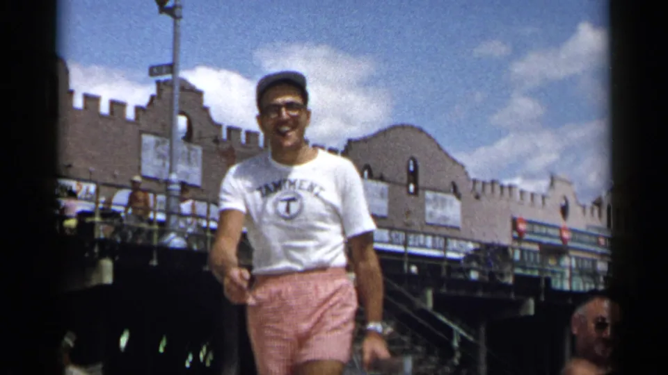 Smiling adult man walks toward the camera on a 1954 New York boardwalk, with signs, a stepped facade, and seated beachgoers in the background.