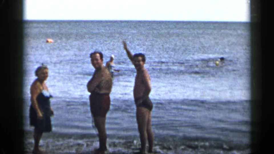 1954 New York 8mm home-movie frame showing three adults at the beach, one waving beside two others in shallow water.