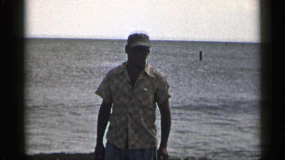 1954 New York beach frame showing a man in a cap and patterned shirt near the shoreline, backlit against open water.