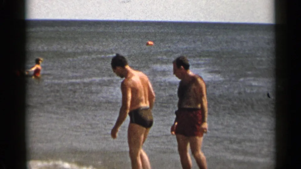 1954 New York 8mm home-movie frame showing two men in swim trunks walking along shallow surf with swimmers offshore.