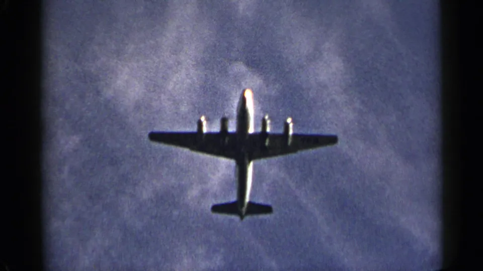 Large propeller aircraft filmed from below in a 1954 New York home-movie frame with blue sky and clouds.