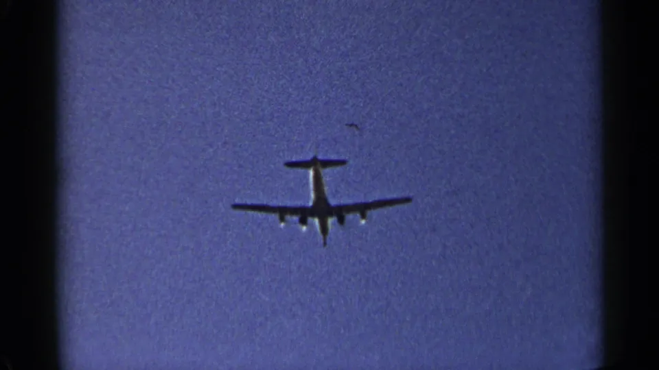 Distant 1954 New York home-movie frame of a propeller aircraft receding in a clear blue sky.