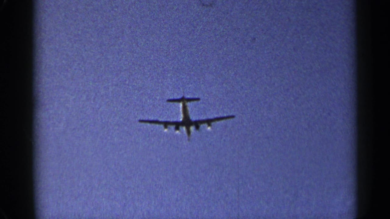 Small airplane silhouette flying across a blue sky with dark 8mm frame edges.