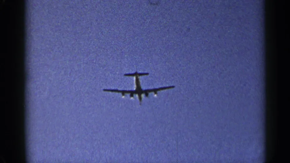 A small airplane appears in silhouette against a deep blue sky in 1954 8mm home-movie footage from New York.