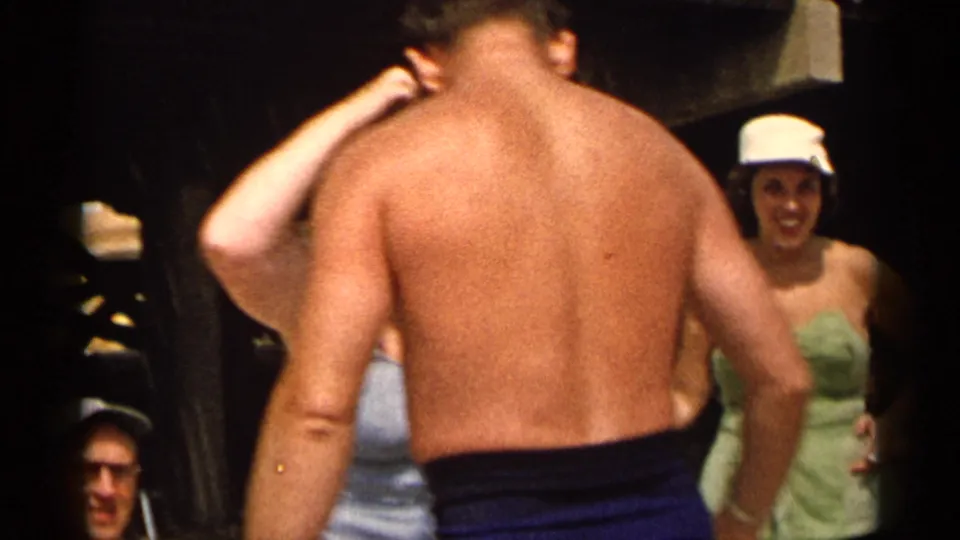 Adults in swimsuits gather casually outdoors, including a shirtless man in blue trunks and a woman in a light cap.