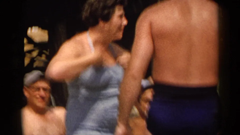 A woman in a light swimsuit appears mid-motion beside a shirtless man, with seated adults visible in the background.