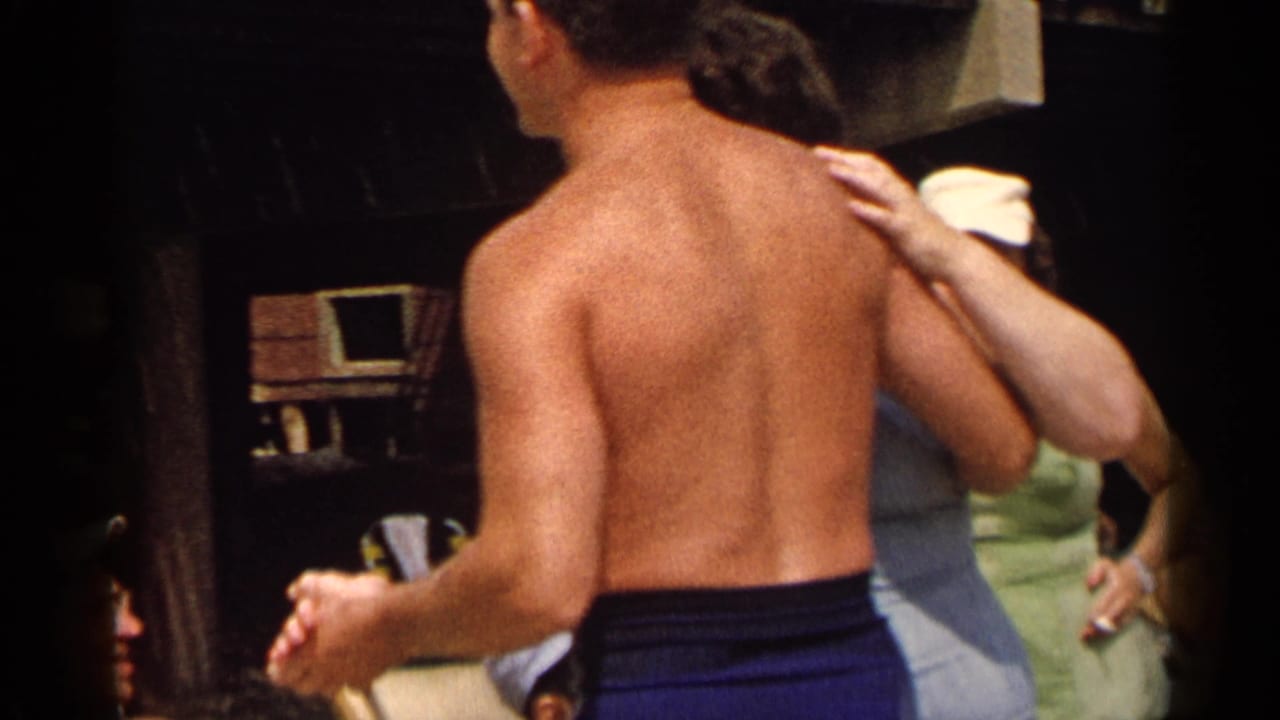 Shirtless man in blue trunks turns near a woman whose hand rests on his shoulder.