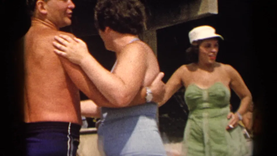 1954 New York home-movie frame of a dancing couple in swimwear, watched by a woman in a green strapless swimsuit and white cap.