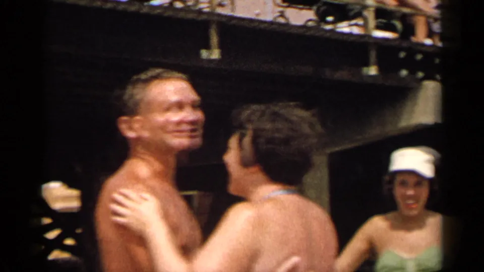 1954 New York home-movie frame of a smiling shirtless man dancing with a woman in a light blue swimsuit at an outdoor party.