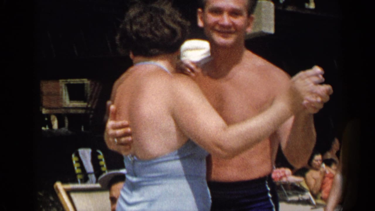 Shirtless man and woman in a light blue swimsuit dance closely on sand.