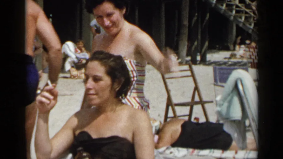 A seated woman smokes beside other adults in swimwear during a 1954 New York beach-party scene.