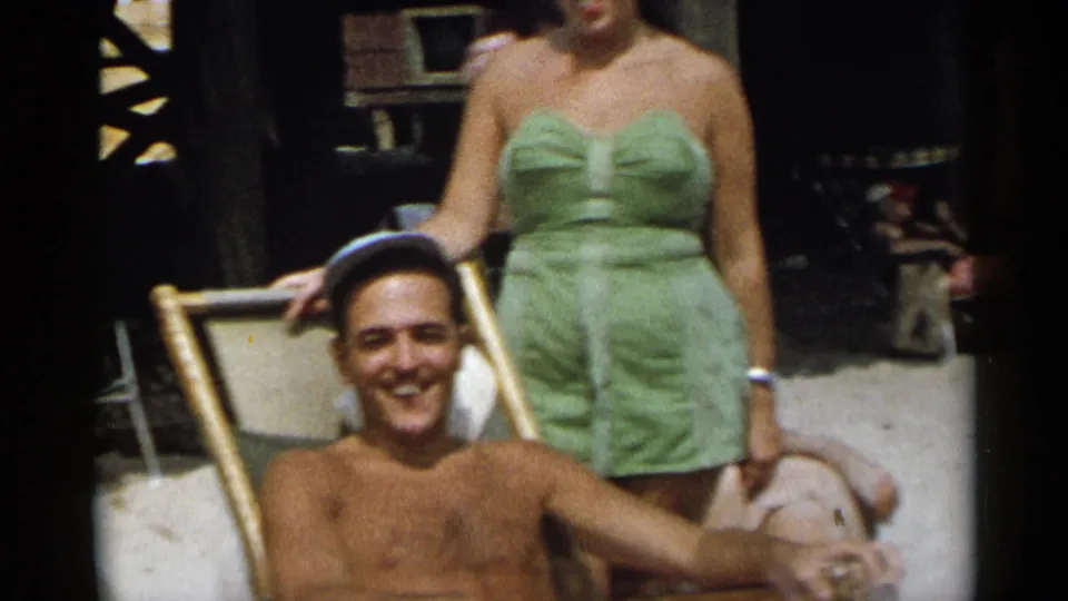 A smiling man seated on a beach chair poses near a woman in a green swimsuit in 1954 New York home-movie footage.
