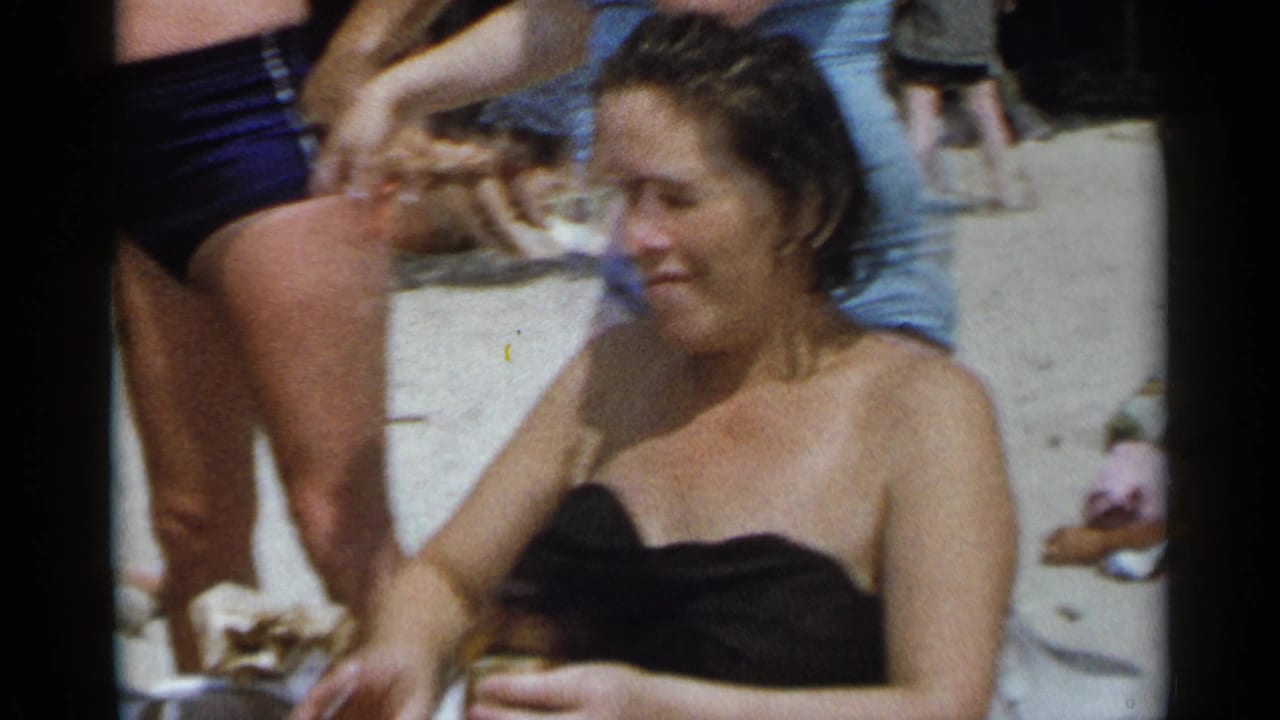 Woman in a black swimsuit sits among other beachgoers and chairs.